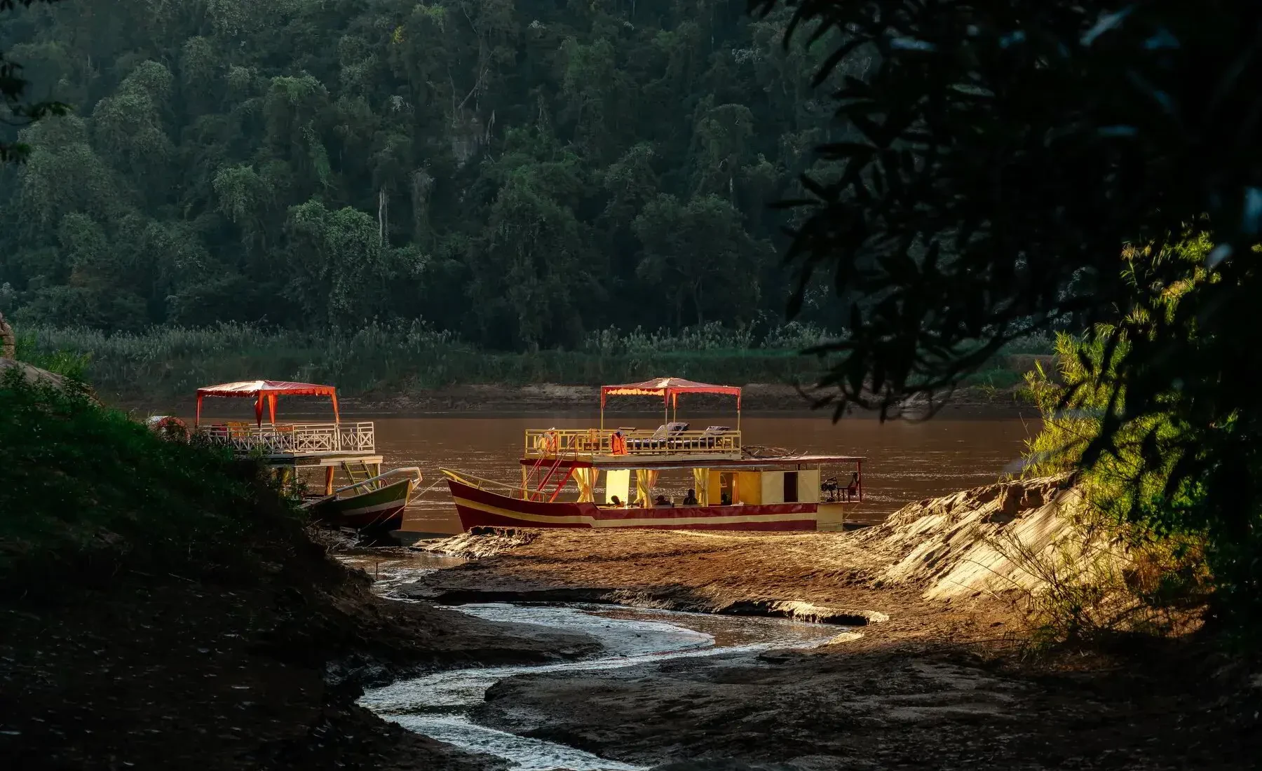 The Tsiribihina river in late afternoon light, with red and yellow river boats moored on a sunlit sandbank below a forested ridge